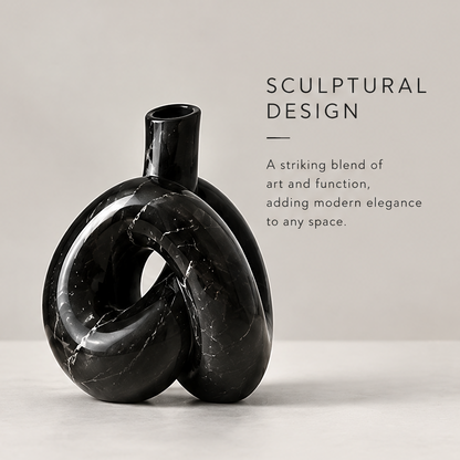 Black marble knot sculptural vase with white flower stems displayed on a console table beside stacked books and a round mirror